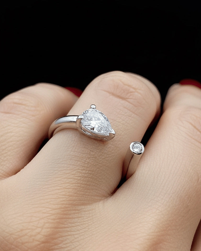 A silver ring with a pear-cut clear gemstone is shown worn on a finger, alongside a smaller matching silver ring with a round clear gemstone, against a black background.